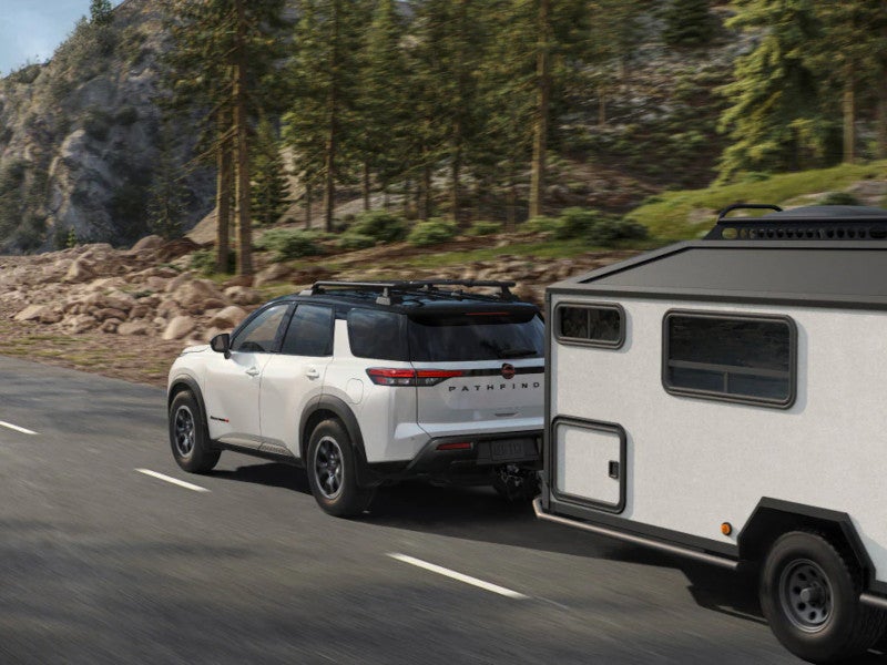 White 2026 Nissan Pathfinder with a black roof and roof rack towing a camper trailer on a mountain road