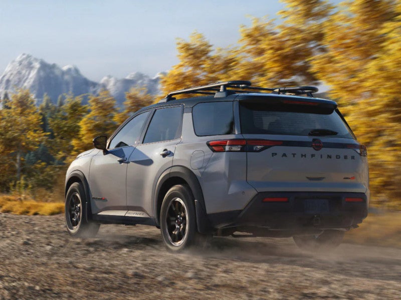 Gray 2026 Nissan Pathfinder with a roof rack driving on a dirt trail past autumn trees and snowy mountains
