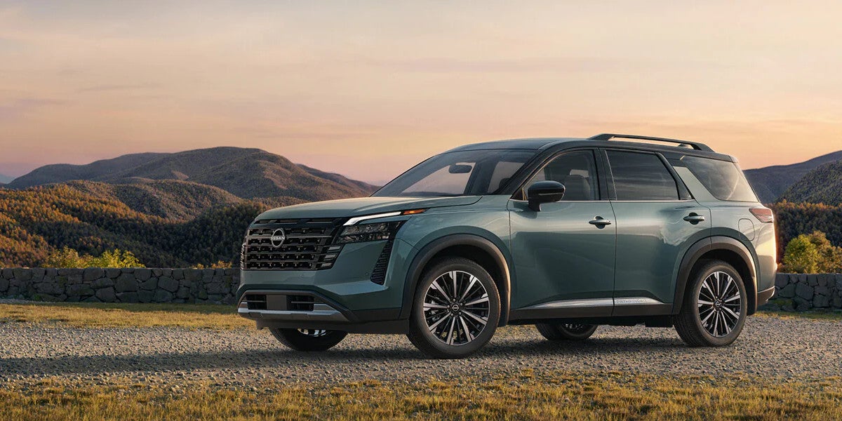 2026 Nissan Pathfinder in a dark green finish parked on a gravel overlook with mountains and a sunset sky behind it