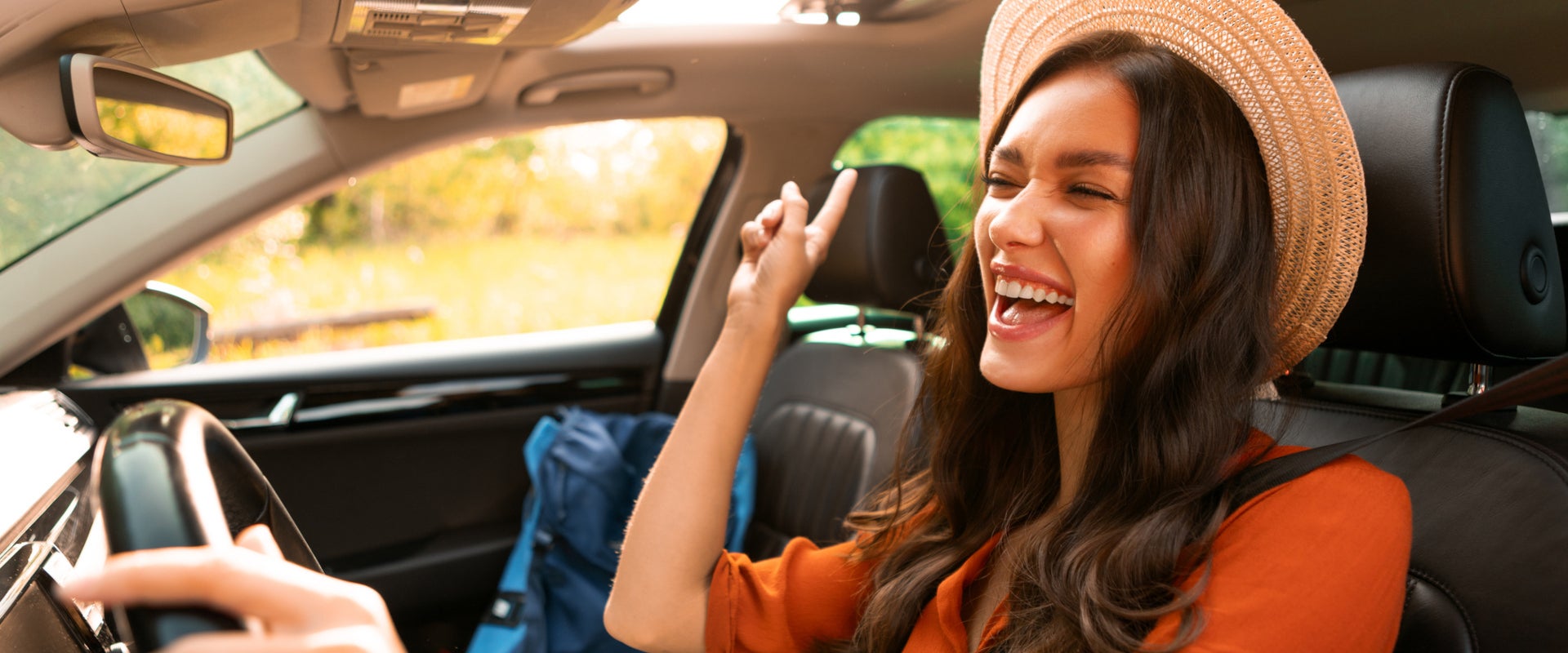 Woman wearing a hat smiles and points upward while sitting in the driver's seat of a car with sunlight streaming in.