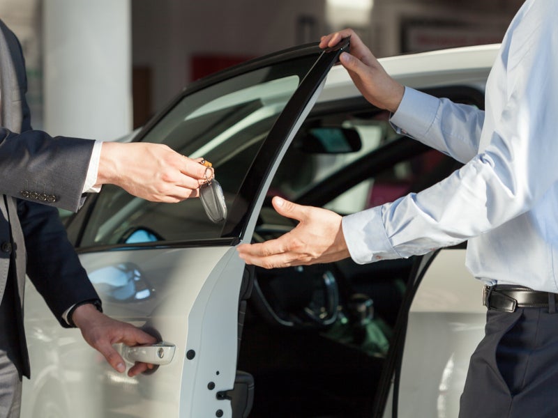 Two people stand by an open car door; one hands over a set of car keys to the other.
